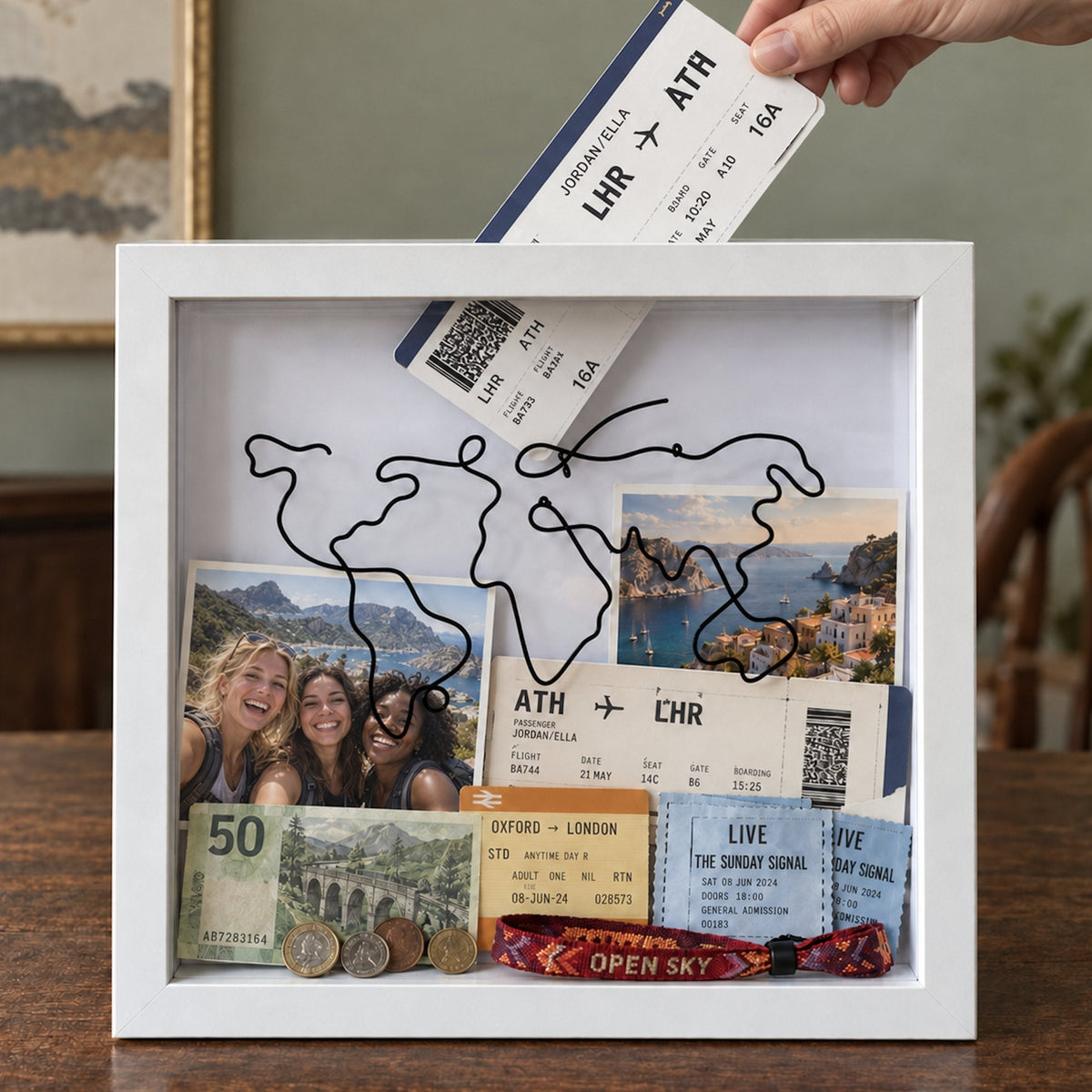 A hand posting a Heathrow to Athens boarding pass through the top slot of a white Wandersake Memory Box filled with photos, tickets and coins