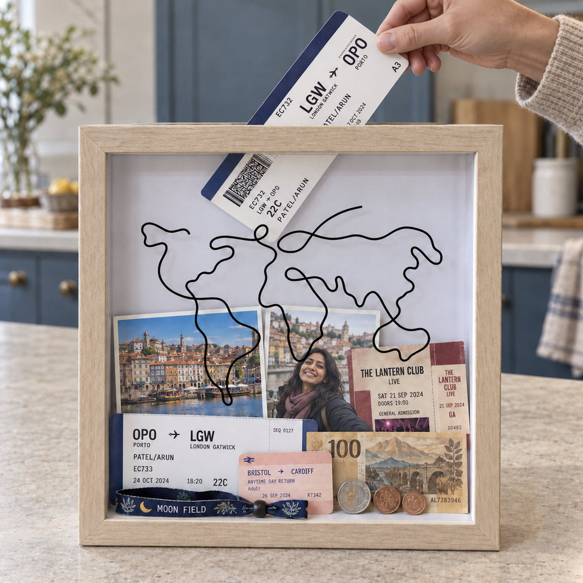 Hand posting a boarding pass into the top slot of a natural wood Wandersake Memory Box on a kitchen worktop, already holding gig tickets and photos from Porto